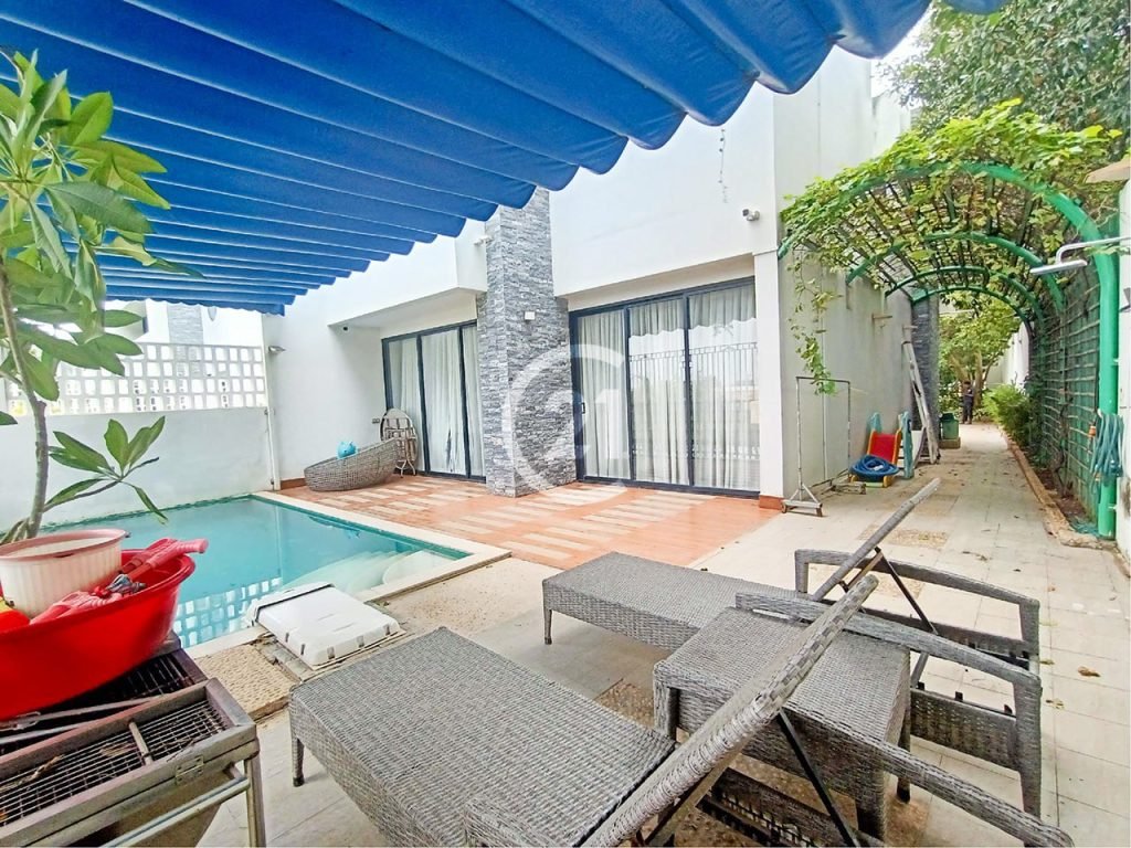 A modern outdoor patio in a twin villa features a small pool, lounge chairs, potted plants, and a blue awning. A walkway with green vines on an arch leads past the pool and house. Glass doors open from the villa onto the patio.