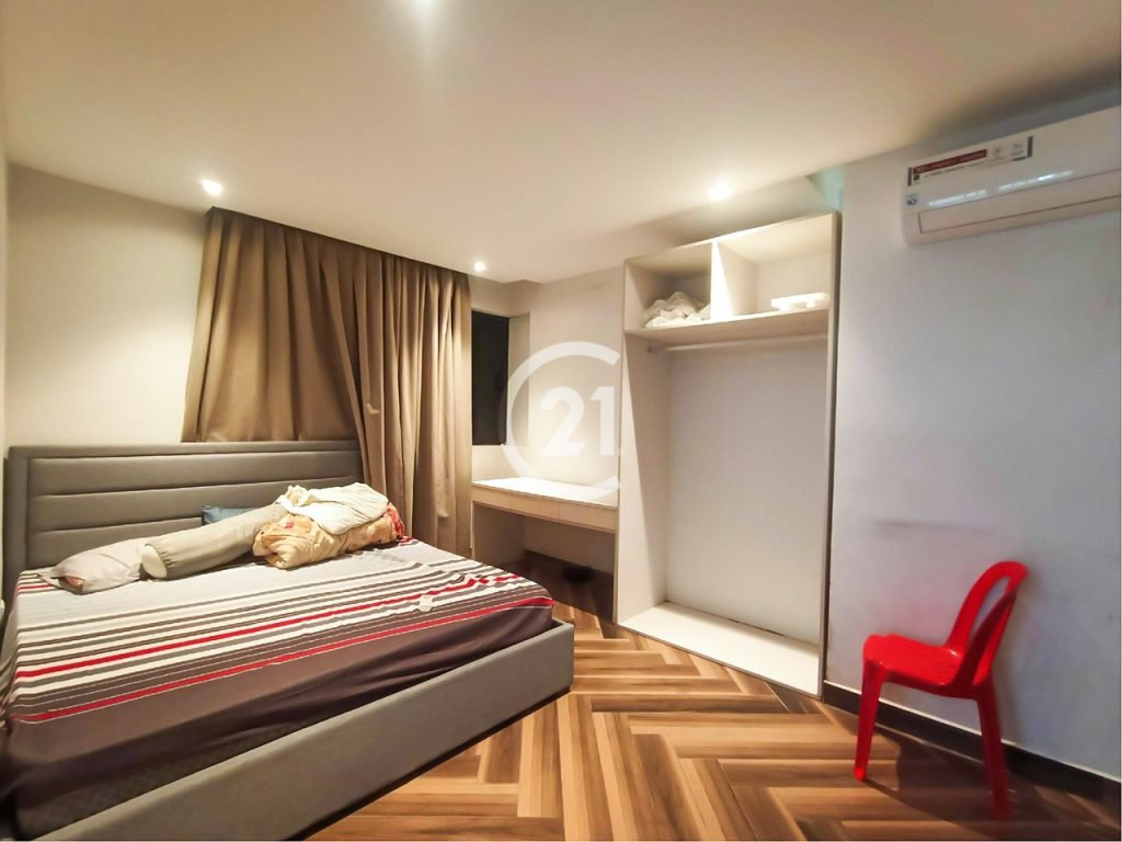 A modern twin villa bedroom with a grey upholstered bed, striped bedding, brown curtains, open closet with shelves, wall-mounted air conditioner, red plastic chair, and wood-patterned floor. The room is brightly lit.