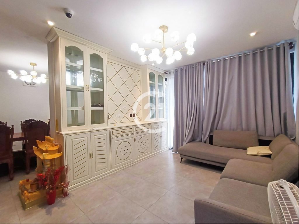 A modern twin villa living room with gray tile flooring, light gray sofas, a large white cabinet with glass doors, and gray curtains. A gold chandelier shines above, while a wooden dining area and a small golden altar complete the space.