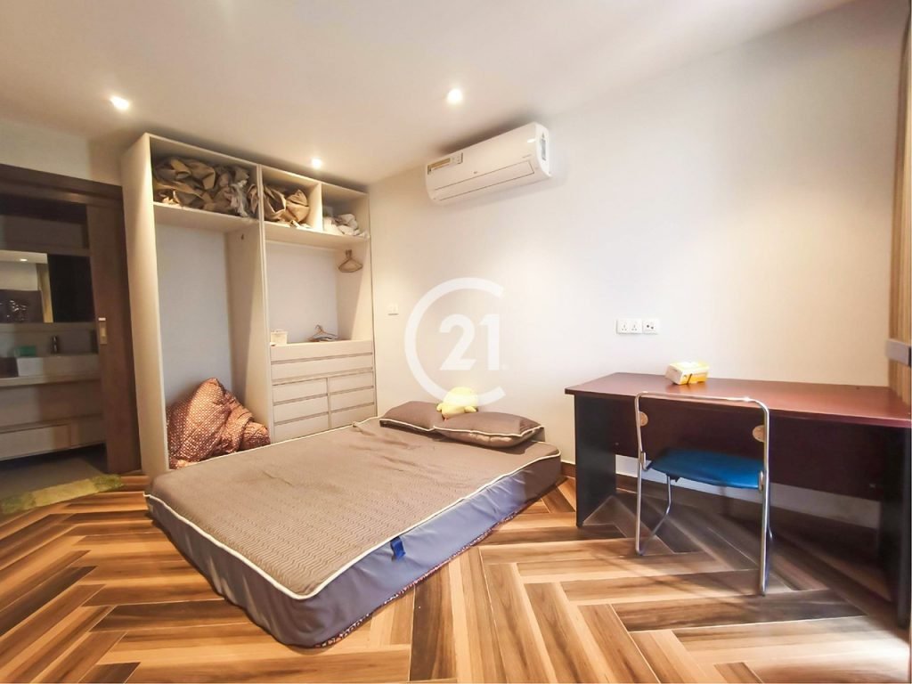 Minimalist bedroom in a twin villa with a mattress on the wooden floor, open shelves and drawers, air conditioner, brown desk with a chair, and soft lighting. A bathroom is visible through an open door.