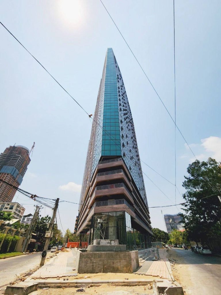 A tall, modern, triangular glass building stands at a street intersection under a bright, sunny sky, with construction materials and wires visible in the foreground and other buildings in the background.
