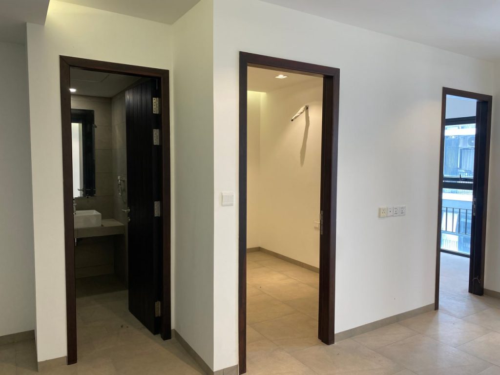A bright, empty room with beige tiled flooring. Two open doorways are visible: one leads to a bathroom with a sink; the other to a smaller room. Large windows on the right bring in natural light.
