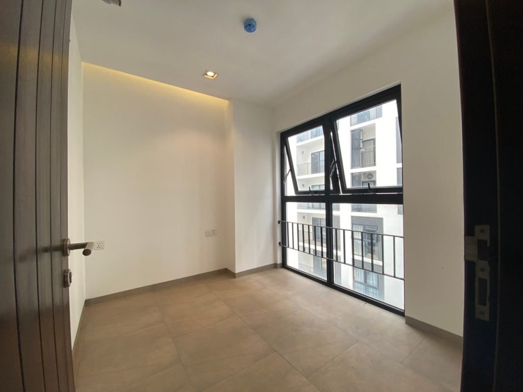 A small, empty room with tiled flooring, white walls, and a large window with black frames and a railing, letting in natural light and showing a view of adjacent buildings.