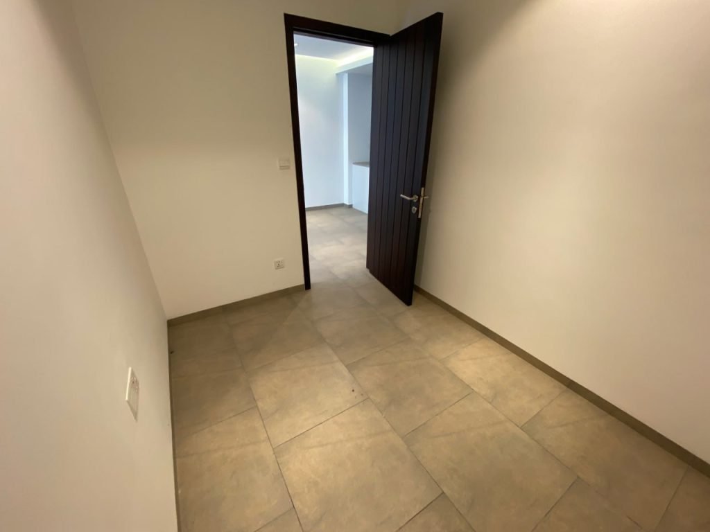 A small empty room with beige tiled floor, white walls, and an open dark wooden door leading to a brighter adjoining space.