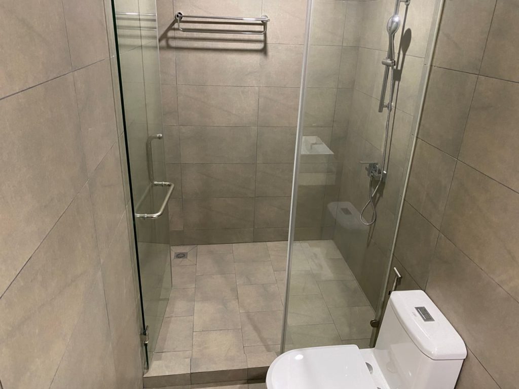 Modern bathroom with beige tiled walls and floor, featuring a glass-enclosed walk-in shower and a white toilet. A metal towel rack is mounted on the wall inside the shower area.