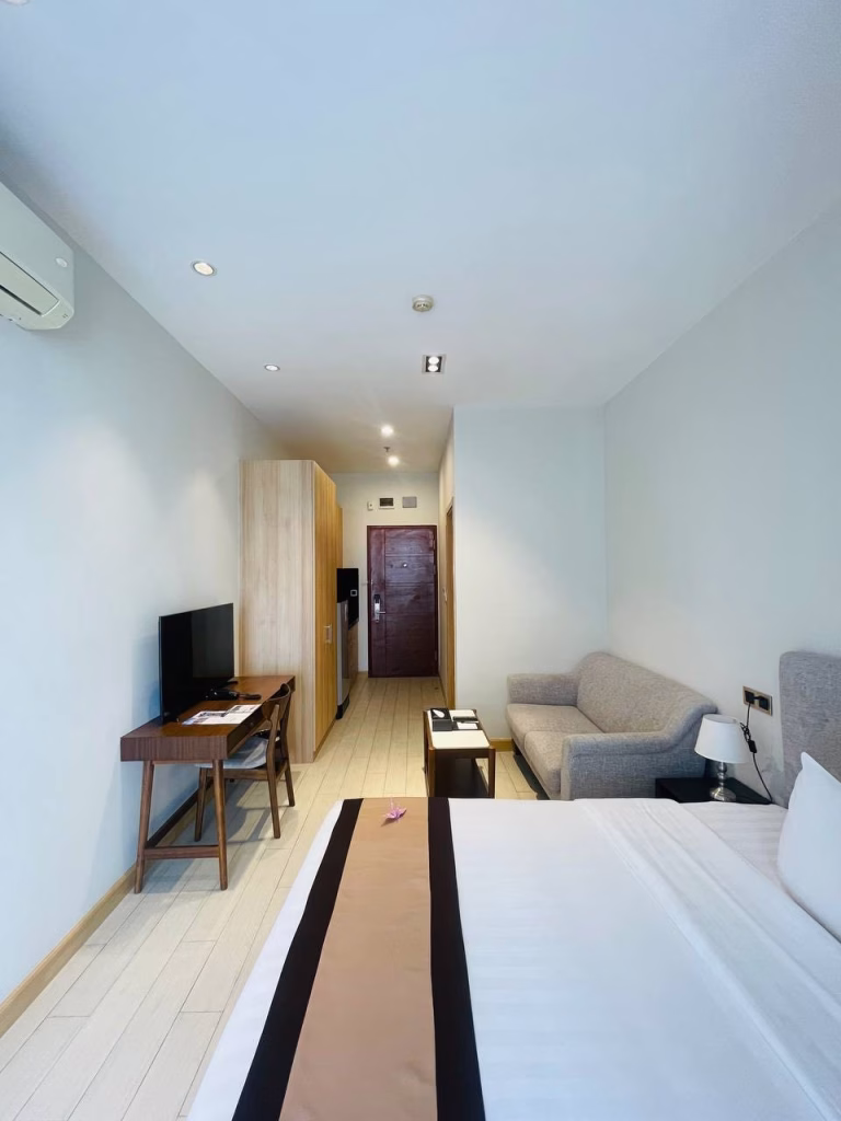 A modern studio for sale featuring a neatly made bed, gray sofa, wooden desk with chair and TV, wardrobe, and a door at the far end. The room boasts white walls and light-colored flooring for a bright, welcoming feel.