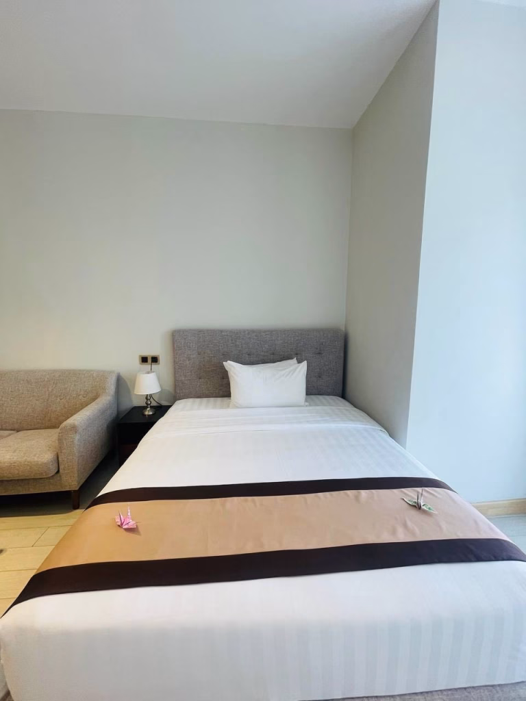 A neatly made single bed with a gray upholstered headboard, white bedding, and a tan and brown runner highlights this inviting studio for sale. Two origami cranes rest on the bed, with a beige armchair and small bedside table nearby.