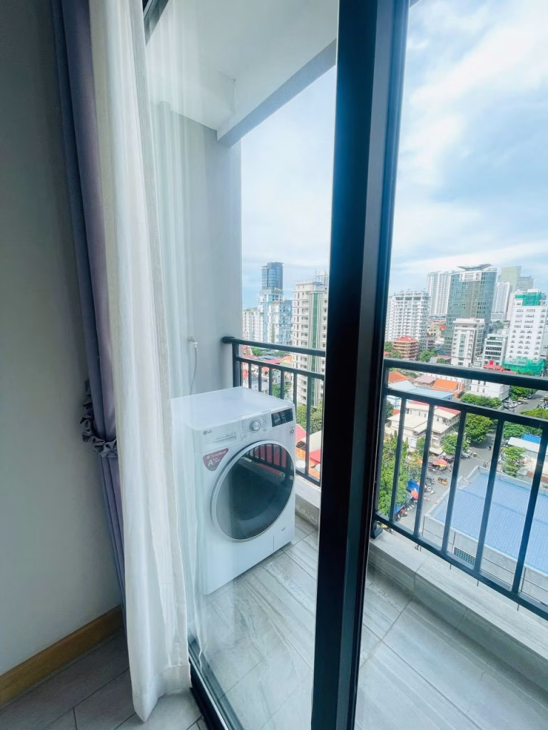 A white washing machine stands on a small balcony with glass railing, overlooking a cityscape—perfect for a studio for sale—seen through a sliding glass door with sheer curtains and partly cloudy sky beyond.