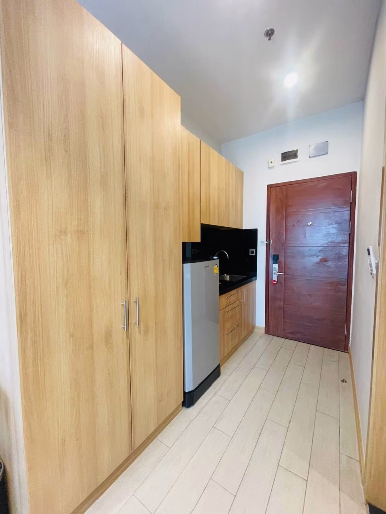 A compact kitchen with light wood cabinets, a small stainless steel refrigerator, a sink, and black backsplash. This studio for sale features white tile flooring, minimal decor, and a wooden entrance door for a modern touch.