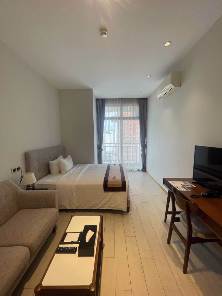 A modern studio for sale featuring a double bed, light gray sofa, wooden desk with a TV, and a coffee table. A large window with sheer curtains lets in natural light, and an air conditioner is mounted on the wall.